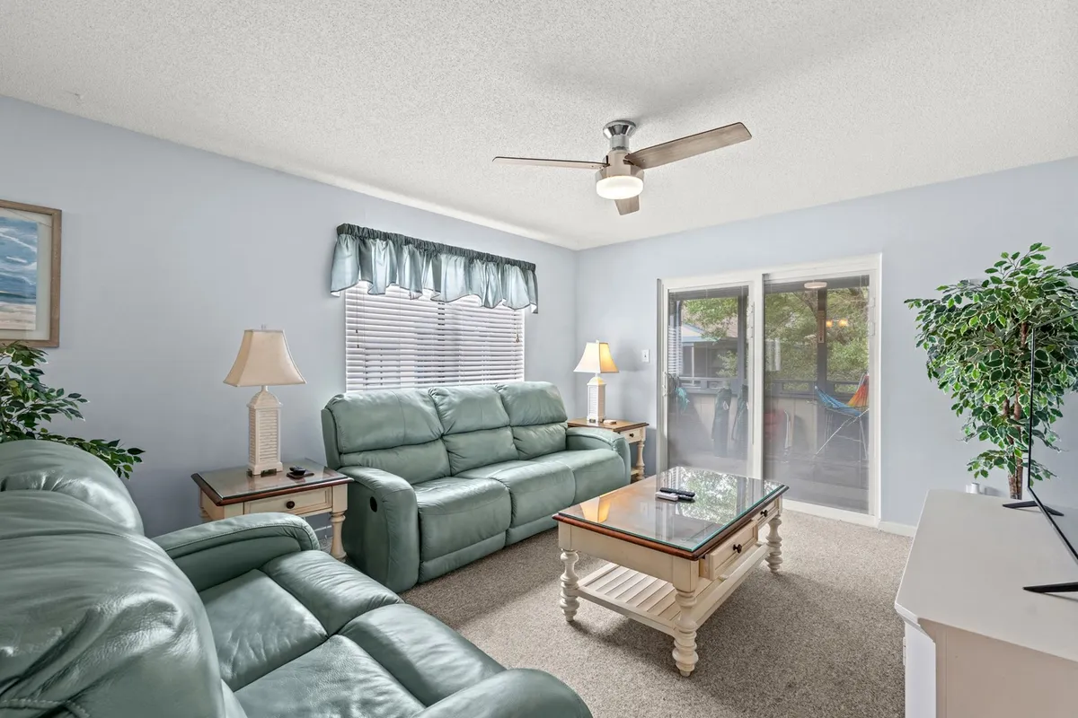 The image showcases the spacious, sunlit living room of the condo with plush green leather sofas arranged around a sleek coffee table. Large windows with soft curtains invite natural light, while decorative plants add a fresh, welcoming touch. The room exudes comfort and is ideal for gathering with family or friends for relaxing, reading, or movie nights.