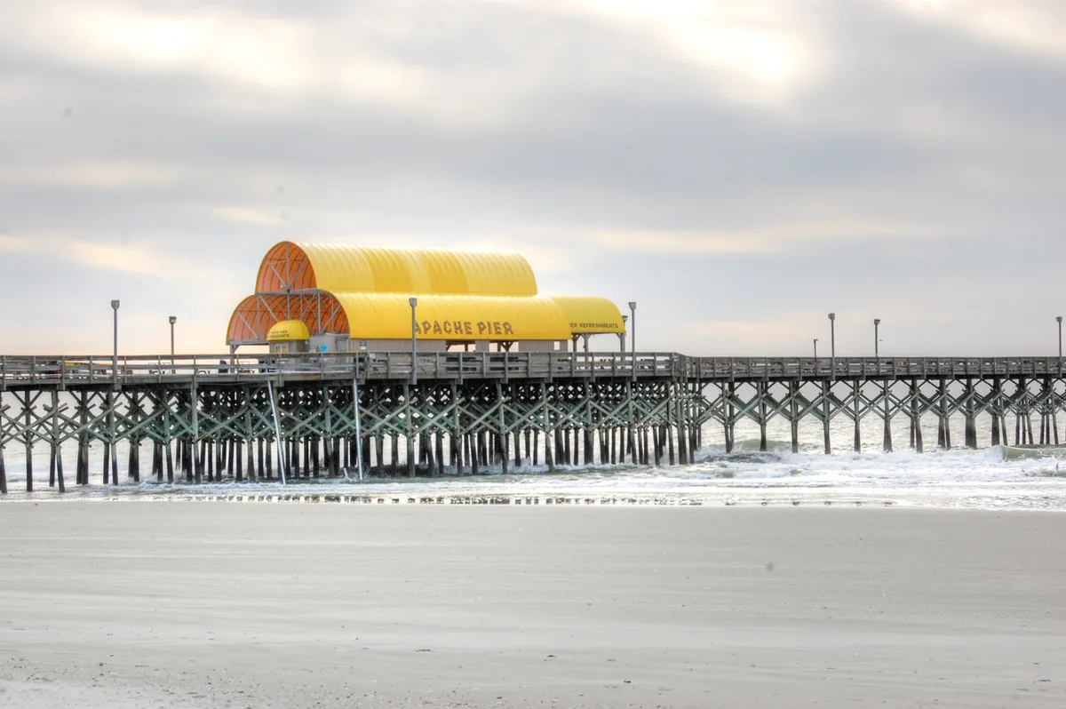This exterior image features the iconic Apache Pier stretching into the ocean, with gentle waves lapping below. The pier building stands as a local landmark, offering a calm and serene setting perfect for long walks or moments of quiet reflection, even during the off-season.