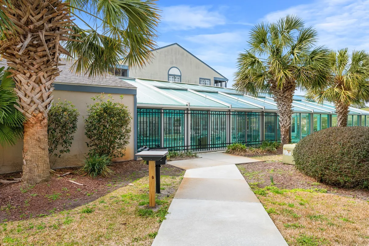 An exterior view of the Summertree Village property, highlighting the landscaped path lined with palm trees and the greenhouse structure. The scene conveys a peaceful, garden-like setting ideal for morning strolls, enjoying the natural surroundings, or savoring quiet coffee on the porch.