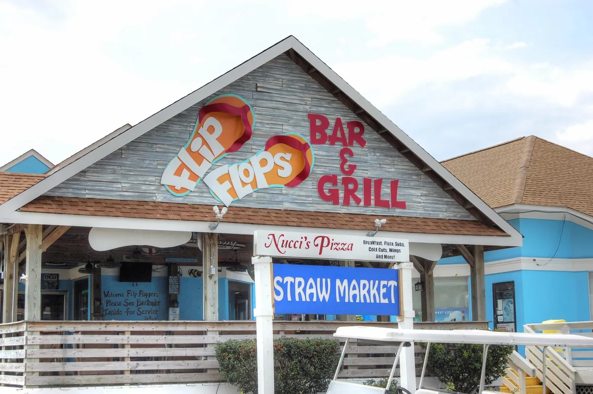 This exterior scene spotlights the lively signs for Flip Flops Bar & Grill, Nucci's Pizza, and the Straw Market, with an inviting, casual atmosphere. The scene hints at nearby dining, social spots, and a welcoming neighborhood vibe for long-term guests.