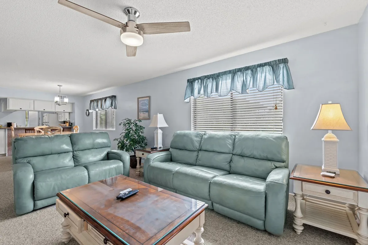 The image features a comfortable living room with green leather sofas arranged around a wooden coffee table. Lamps cast a warm glow, and a large window with blue curtains lets in abundant natural light, creating a bright and inviting space ideal for relaxation and social activities.