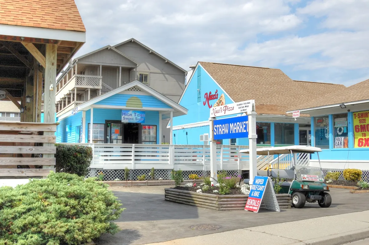 A welcoming exterior view of local shops and restaurants, including a pizza place and a smoke shop, along with a golf cart parked nearby. The scene conveys the ease of exploring local dining, shopping, and leisure options on a sunny day.