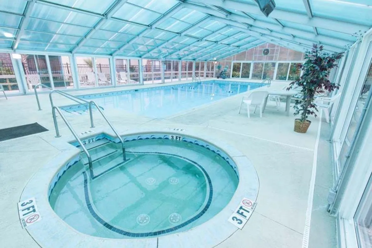 This interior image showcases a tranquil indoor swimming pool with a hot tub and surrounding seating area. Sunlight streams in through large windows, creating a serene atmosphere perfect for relaxation and socializing. The pool is inviting, while the hot tub offers opportunities for soothing soaks after a swim.