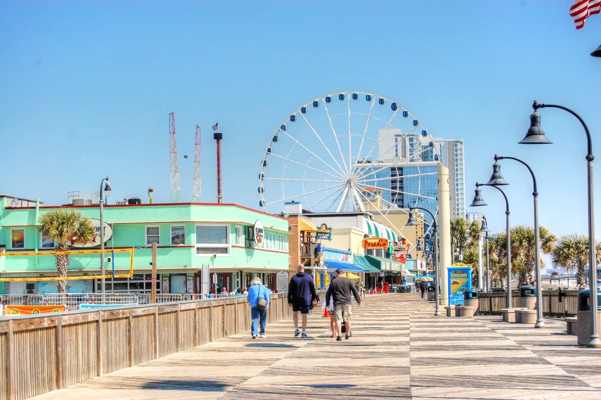 Set in early evening light, this exterior scene showcases the ferris wheel, colorful storefronts, and people exploring the boardwalk. The setting sun adds a magical, inviting atmosphere, emphasizing the social and playful vacation vibe.