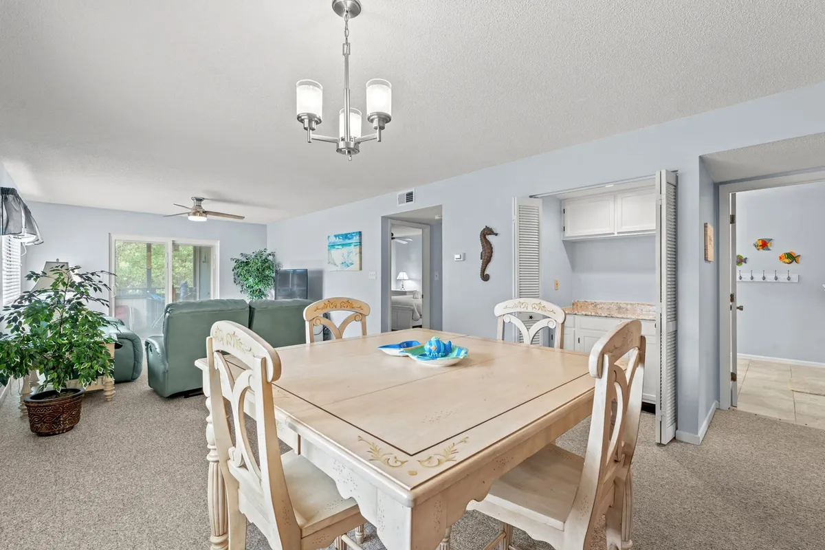 This scene looks into the open-concept dining and living area, with a dining table situated near a large window and a green sofa in the background. A hanging light fixture and decorative fish add coastal charm, while the overall space is bathed in daylight, emphasizing the seamless flow between dining and relaxing.