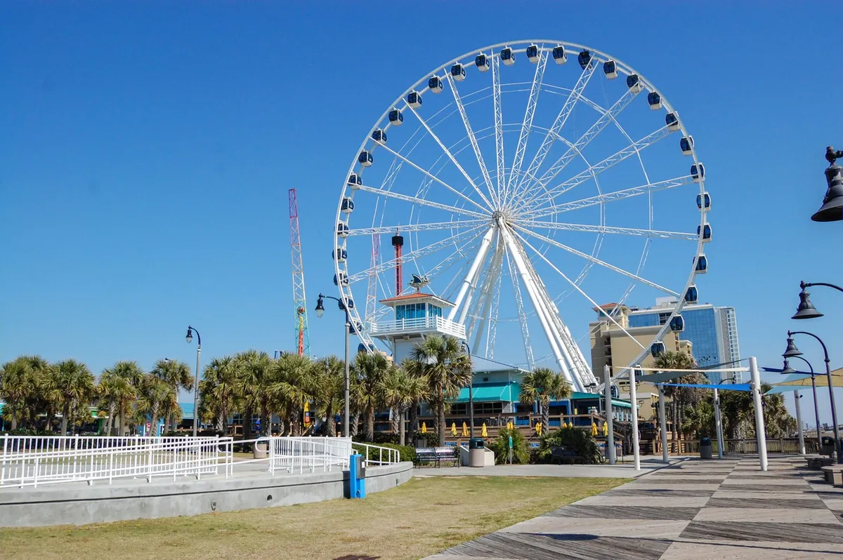 This vibrant exterior image features the iconic Myrtle Beach ferris wheel, the bustling boardwalk, palm trees, and an amusement center. People are visible strolling, enjoying rides, and sampling treats, capturing the lively energy and range of activities available at local attractions.
