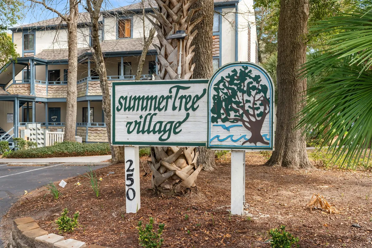 The welcoming exterior of Summer Tree Village, featuring the community's sign, mature trees, and inviting architecture. The scene suggests a peaceful, family-friendly neighborhood with lush landscaping and communal spaces for gatherings or strolls.