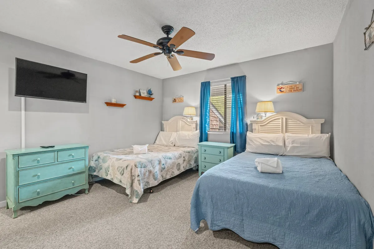 A comfortable bedroom featuring two double beds with light bedding, a turquoise dresser, cheerful blue curtains, and decorative wall shelves. The space appears cozy and is ideal for families or groups, encouraging relaxation or shared moments.
